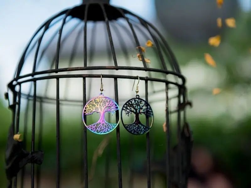 Metall-Ohrringe in Baum-Optik von Martina als Goldschmiedin. Sie glänzen in Regenbogen-Farben und hängen an einem Käfig mit schwarzen Gitterstäben, der sich im Freien befindet.