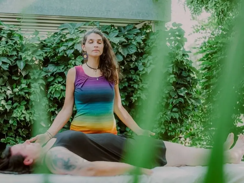 Martina steht mit geschlossenen Augen vor einer liegenden Person und hat ihre offenen Arme und Hände über ihr ausgebreitet, während sie bei Reiki bzw. Seelenkommunikation die Energie fließen lässt.