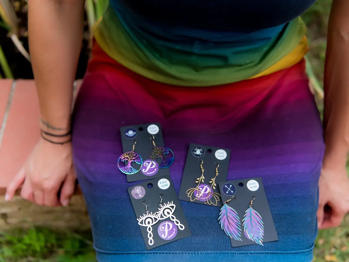 Energieschmuck Ohrringe verschiedene Energetisierte Edelstahl-Ohrringe in verschiedernen Designs und schimmern in Regenbogen-Farben liegen im Schoß einer Frau, die ein farbenfrohes Kleid trägt.